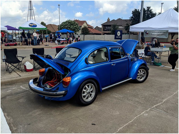 A blue car parked in a parking lot with people and tentsDescription automatically generated with low confidence